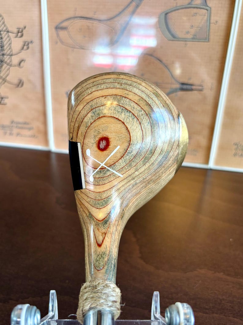 Puede incluir: Un palo de golf de madera vintage con un patr&oacute;n de vetas de madera marr&oacute;n claro y beige. La cabeza del palo presenta un acento negro y un dise&ntilde;o de tee de golf cruzado blanco. El agarre est&aacute; envuelto con hilo natural.