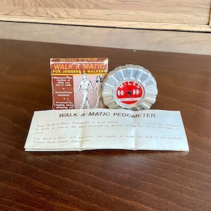 May include: A silver and red pedometer with the text "WALK-A-MATIC" and "MILES" on it. The pedometer is packaged in a box with the text "WALK-A-MATIC FOR JOGGERS & WALKERS".