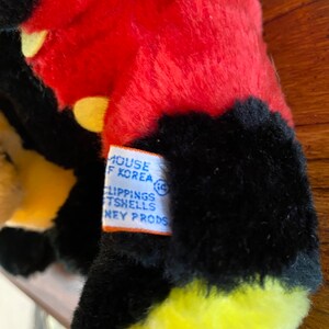 May include: Close-up of a plush toy with red, black, and yellow fur. The toy has a tag that reads "MOUSE OF KOREA" and "CLIPPINGS TSHELLS DISNEY PRODS."