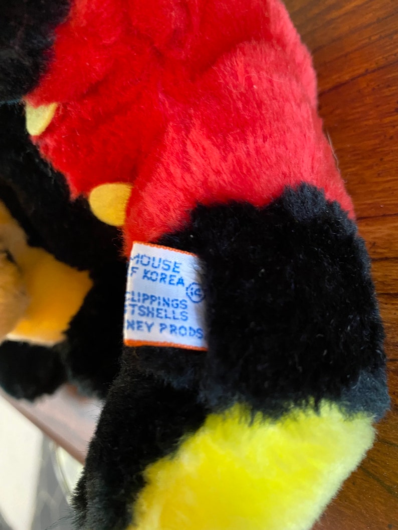 May include: Close-up of a plush toy with red, black, and yellow fur. The toy has a tag that reads "MOUSE OF KOREA" and "CLIPPINGS TSHELLS DISNEY PRODS."