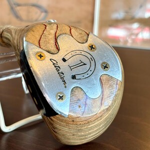 Puede incluir: Palo de golf de madera vintage con una cara plateada. La cara presenta el n&uacute;mero "1" dentro de una herradura, la palabra "Citation" y cuatro tornillos dorados. La cabeza del palo tiene un patr&oacute;n de vetas de madera clara con acentos rojos.