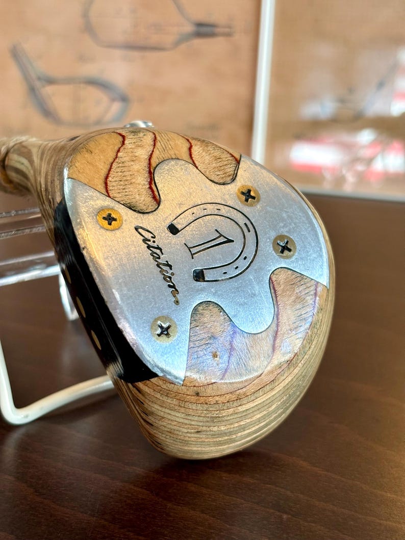 Puede incluir: Palo de golf de madera vintage con una cara plateada. La cara presenta el n&uacute;mero "1" dentro de una herradura, la palabra "Citation" y cuatro tornillos dorados. La cabeza del palo tiene un patr&oacute;n de vetas de madera clara con acentos rojos.
