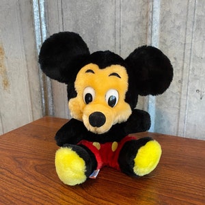 May include: A plush Mickey Mouse toy with black ears, a yellow face, and a black nose. The toy is wearing red shorts with yellow buttons and yellow feet. The toy is sitting on a wooden surface.