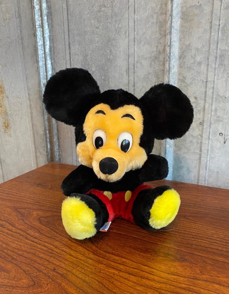 May include: A plush Mickey Mouse toy with black ears, a yellow face, and a black nose. The toy is wearing red shorts with yellow buttons and yellow feet. The toy is sitting on a wooden surface.