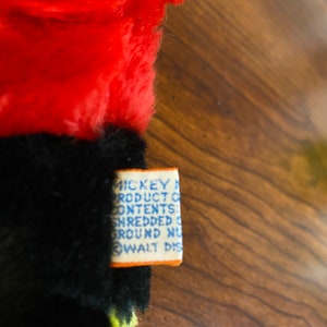 May include: Close-up of a vintage plush toy with red, black, and yellow fur. A rectangular tag with blue text and an orange border is attached to the toy. The tag reads "MICKEY MOUSE PRODUCT CONTENTS: SHREDDED GROUND NU... WALT DIS..."