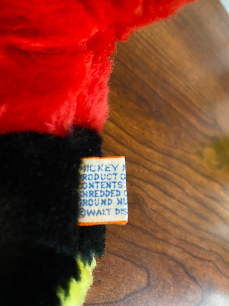 May include: Close-up of a vintage plush toy with red, black, and yellow fur. A rectangular tag with blue text and an orange border is attached to the toy. The tag reads "MICKEY MOUSE PRODUCT CONTENTS: SHREDDED GROUND NU... WALT DIS..."