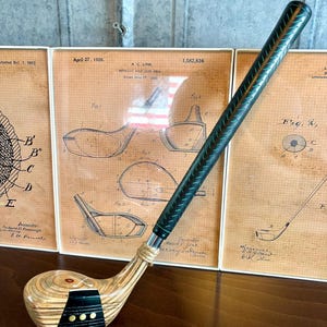 Puede incluir: Un palo de golf vintage con una cabeza de madera y un agarre verde oscuro. El palo se muestra frente a dibujos de patentes enmarcados. La cabeza tiene una franja negra con tres puntos dorados. La fecha de la patente es el 27 de abril de 1926.