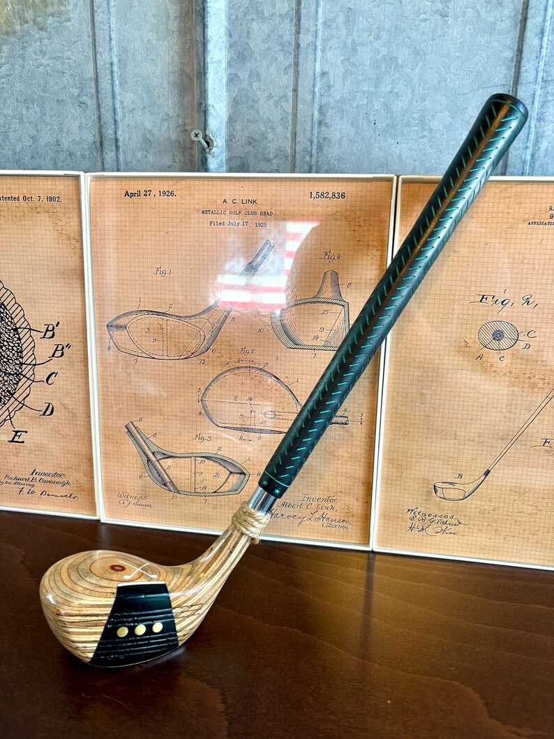 Puede incluir: Un palo de golf vintage con una cabeza de madera y un agarre verde oscuro. El palo se muestra frente a dibujos de patentes enmarcados. La cabeza tiene una franja negra con tres puntos dorados. La fecha de la patente es el 27 de abril de 1926.