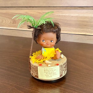 May include: A musical toy featuring a brown-skinned doll wearing a yellow dress and a lei, sitting on a beach scene with a palm tree. The doll is playing a ukulele and the text on the base reads "Waikiki Kimo Basking in the sun Round he goes the music plays "Aloha" he says to everyone"