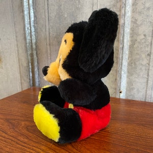May include: A plush Mickey Mouse toy, featuring black fur, a yellow face, and red shorts. The toy has large black ears and yellow feet. The toy is sitting on a wooden surface.