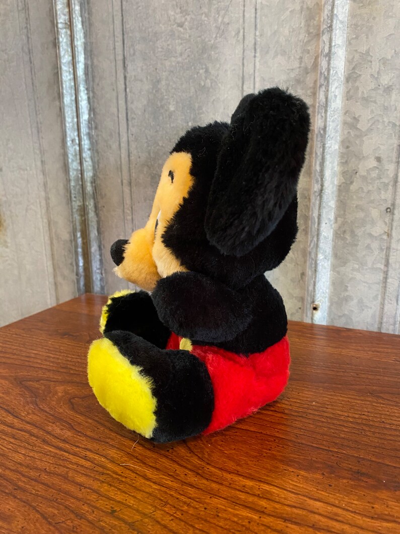 May include: A plush Mickey Mouse toy, featuring black fur, a yellow face, and red shorts. The toy has large black ears and yellow feet. The toy is sitting on a wooden surface.