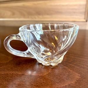 May include: A clear glass teacup with a decorative, swirled design. The cup has a rounded base and a handle. The teacup is sitting on a dark brown surface.