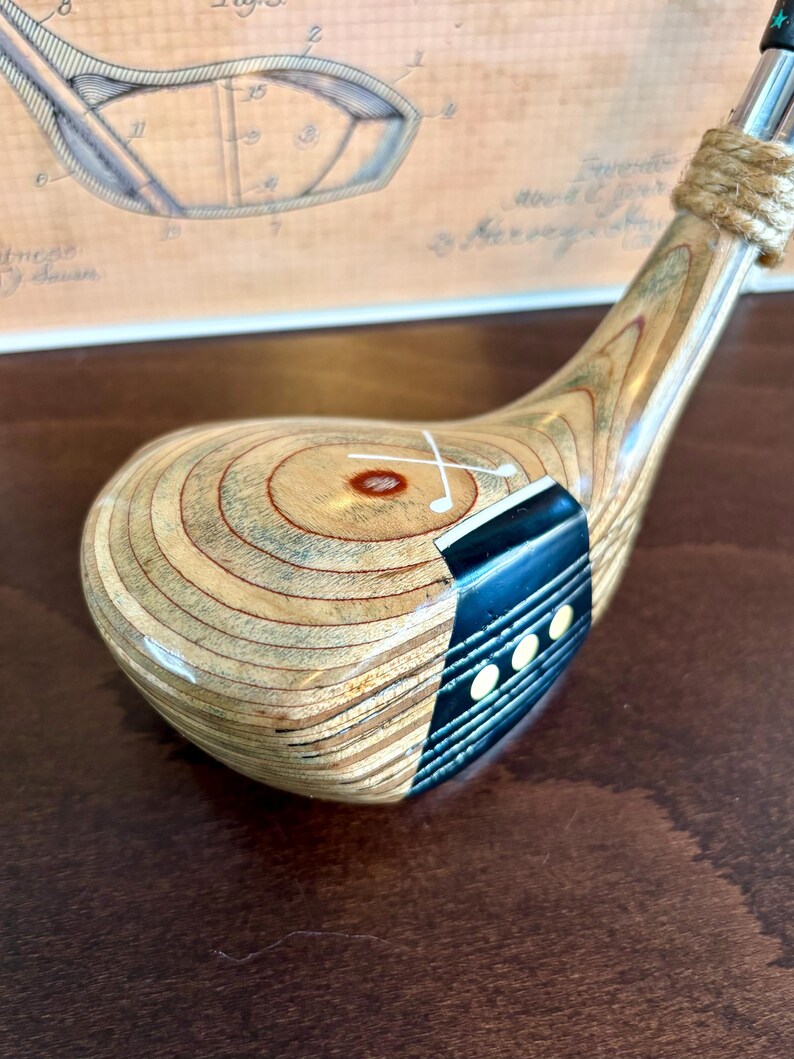 Puede incluir: Un palo de golf de madera vintage con una cabeza marr&oacute;n claro y un acento azul oscuro. El eje del palo es plateado y el agarre est&aacute; envuelto con hilo. La cabeza presenta una marca blanca y tres peque&ntilde;os puntos blancos.