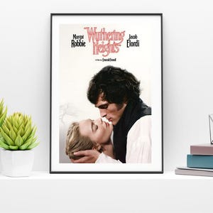 Puede incluir: Un p&oacute;ster enmarcado de la pel&iacute;cula "Cumbres Borrascosas" con Margot Robbie y Jacob Elordi. El p&oacute;ster muestra un primer plano de los actores bes&aacute;ndose. Dos plantas suculentas verdes en macetas blancas y una pila de libros en una estanter&iacute;a blanca.