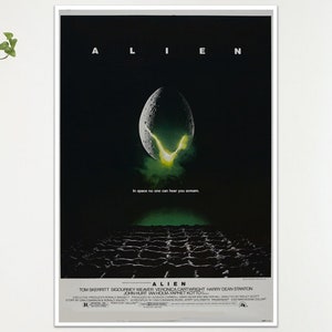 May include: A framed movie poster for the film "Alien." The poster features the title in white capital letters at the top, with an egg-shaped object in the centre emitting green light. The bottom of the poster has the film's credits.