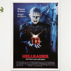 May include: A framed poster for the horror film "Hellraiser". The image shows a character with pins in their head holding a glowing box. The poster's title is in red, with the tagline "He'll tear your soul apart."