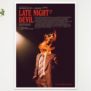 May include: A framed poster for the film "Late Night with the Devil" shows a man in a suit with a flaming head. The poster has a dark background with red and white text, including the film's title.