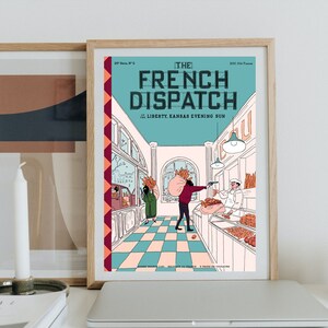 May include: Framed print of "The French Dispatch" magazine cover. The illustration features a bakery scene with figures and text. The print is in a wooden frame, displayed with other decor on a white surface.