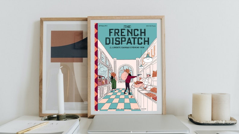 May include: Framed print of "The French Dispatch" magazine cover. The illustration features a bakery scene with figures and text. The print is in a wooden frame, displayed with other decor on a white surface.
