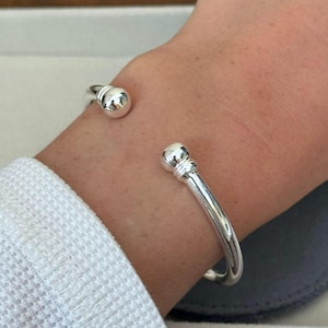 May include: A silver cuff bracelet with a spherical design at each end. The bracelet is worn on a wrist. The metal has a shiny, reflective surface. The cuff is a simple, elegant piece of jewellery. The bracelet's diameter is approximately 6 cm.