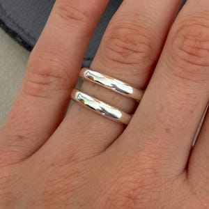 May include: A silver ring with two parallel bands is shown on a hand with pink nail polish. A small, grey, snap-closure pouch with a gold button is in the background. The ring has a simple, modern design.
