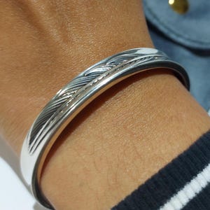May include: A polished silver cuff bracelet with a feather design. The bracelet is worn on a wrist, and the design features detailed lines and curves. A blue pouch with a gold button is in the background.