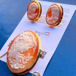 May include: A gold-toned jewelry set featuring cameo designs. The set includes a pendant necklace and matching earrings. The cameos depict a woman's profile in white relief against an orange background. The words "Made in Italy" are visible.