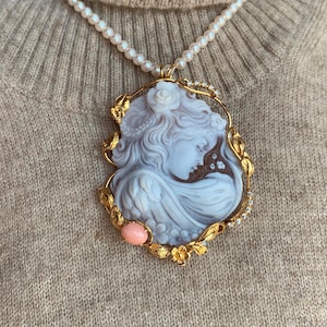 May include: A gold pendant necklace with a cameo featuring a woman's profile. The cameo is set in a gold frame with floral details and a pink stone accent.
