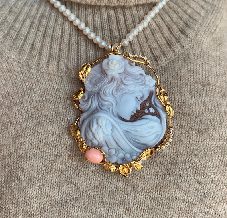 May include: A gold pendant necklace with a cameo featuring a woman's profile. The cameo is set in a gold frame with floral details and a pink stone accent.
