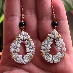 May include: A pair of white carved floral drop earrings with black beads and small clear stones. The earrings are made of shell and have a gold tone finish.
