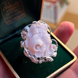 Pode incluir: Um anel de prata com um camafeu esculpido em rosa pálido de um rosto de perfil rodeado por flores e folhas. A banda de metal é acentuada com pequenas pedras claras. O anel é apresentado numa caixa forrada a veludo verde escuro.