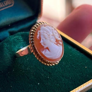 May include: A gold ring with an oval cameo depicting a woman's profile. The cameo is set within a gold frame with a rope-style border. The ring is presented on a green velvet surface.