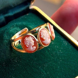 Peut inclure: Une bague dorée ornée de trois camées ovales. Chaque camée présente un relief de profil rose clair sur un fond rouge-orangé plus foncé. La bague possède un anneau ouvert, mettant en valeur les détails sculptés de chaque élément décoratif.