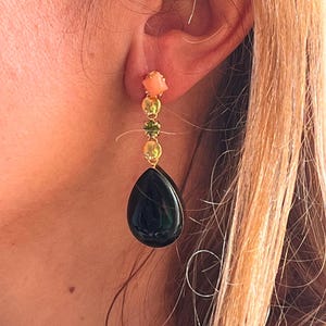 May include: A gold earring featuring a coral square, three gold beads, and a green gem. A large, dark green teardrop stone dangles below. The earring is displayed on a person's ear.