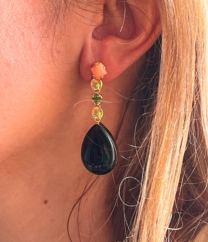 May include: A gold earring featuring a coral square, three gold beads, and a green gem. A large, dark green teardrop stone dangles below. The earring is displayed on a person's ear.