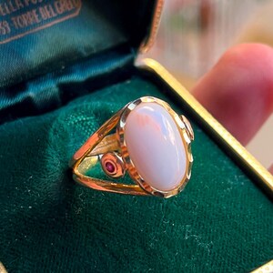 Può includere: Un anello d'oro con una pietra preziosa ovale rosa pallido. L'anello presenta un design delicato con una piccola gemma rossa rotonda sul lato. Presentato in un portagioie di velluto verde.