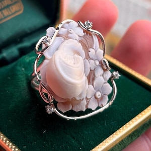 Könnte beinhalten: Nahaufnahme eines Rings mit einem geschnitzten Cameo-Design. Der Ring hat ein silberfarbenes Metallband und einen floralen Rahmen mit kleinen, klaren Edelsteinen. Das Cameo ist hellbeige und der Ring ist vor einem grünen Samthintergrund platziert.