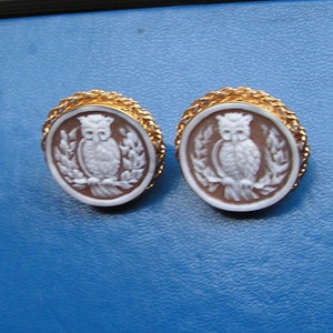May include: A pair of gold tone earrings with a cameo of an owl on each. The owl is white and the cameo is set in a brown background.