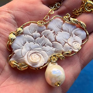 Puede incluir: Collar dorado con un colgante floral blanco tallado, con detalles de rosas y una gota de perla en forma de pera. El colgante est&aacute; enmarcado por hojas doradas y una delicada cadena.