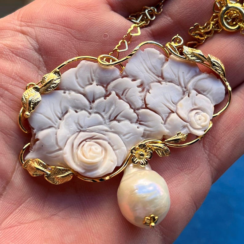 Puede incluir: Collar dorado con un colgante floral blanco tallado, con detalles de rosas y una gota de perla en forma de pera. El colgante est&aacute; enmarcado por hojas doradas y una delicada cadena.