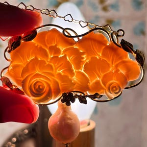 Puede incluir: Un colgante floral naranja con un marco dorado y un peque&ntilde;o dije en forma de pera. El colgante presenta un dise&ntilde;o de rosa detallado. El marco incluye una cadena de peque&ntilde;as formas de coraz&oacute;n.