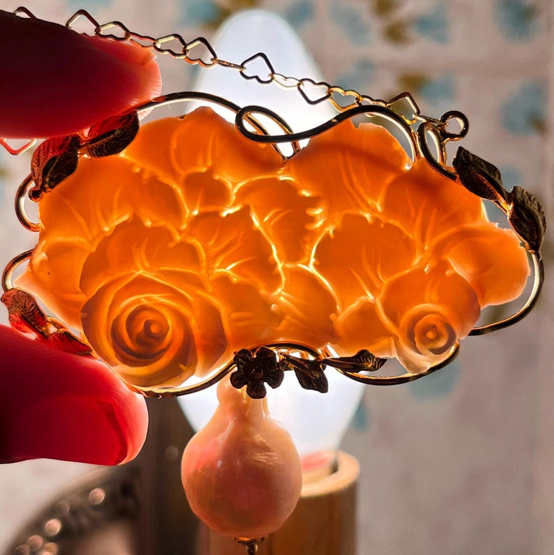 Puede incluir: Un colgante floral naranja con un marco dorado y un peque&ntilde;o dije en forma de pera. El colgante presenta un dise&ntilde;o de rosa detallado. El marco incluye una cadena de peque&ntilde;as formas de coraz&oacute;n.