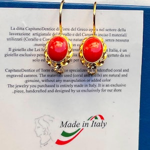 Könnte beinhalten: Ein Paar goldene Ohrhänger mit ovalen, glatten roten Korallen-Cabochons, die in einem dekorativen, gedrehten Golddrahtrahmen gefasst sind. Die Ohrringe sind auf einer weißen Karte mit italienischem und englischem Text sowie einem Made in Italy-Logo abgebildet.