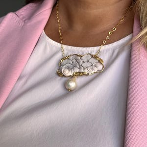 Puede incluir: Un collar dorado con un colgante que representa un dise&ntilde;o floral en tonos grises y blancos. Una gran perla en forma de pera cuelga del colgante. El collar se lleva sobre una camiseta blanca y una blazer rosa.