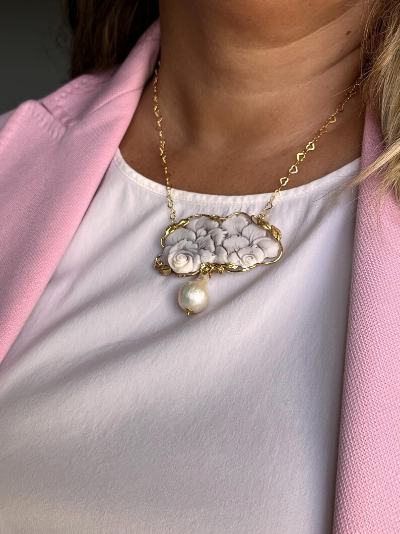 Puede incluir: Un collar dorado con un colgante que representa un dise&ntilde;o floral en tonos grises y blancos. Una gran perla en forma de pera cuelga del colgante. El collar se lleva sobre una camiseta blanca y una blazer rosa.