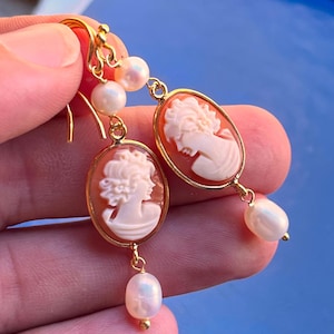 Könnte beinhalten: Goldfarbene Ohrringe mit ovalen Cameo-Anhängern mit weißen Profilporträts. Jeder Ohrring hat eine hängende Perle und kleinere Perlen oben. Die Ohrringe werden von einer Hand gehalten.