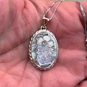 Op de afbeelding: Ovale hangerketting met een zilveren ketting. De hanger heeft een gesneden bloemenontwerp in iriserend parelmoer, gezet in een zilveren frame met een gedraaide touwrand. De ketting wordt getoond tegen een huidkleurige achtergrond.