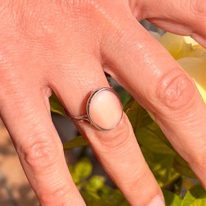 K&ouml;nnte beinhalten: Ein Silberring mit einem gro&szlig;en, ovalen, hellrosa Edelstein. Der Edelstein ist in einem polierten Silberband gefasst. Der Ring wird an einem Finger gezeigt, mit einem weichen Hintergrund.