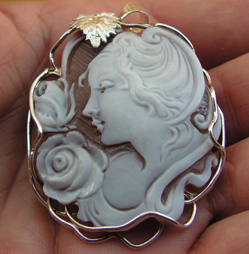 May include: A cameo pendant featuring a woman's profile with a rose, carved from white shell and set in a silver filigree frame.