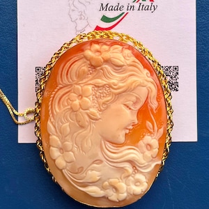 Puede incluir: Colgante de camafeo ovalado con el perfil de una mujer con cabello suelto y detalles florales. El camafeo está engastado en un marco dorado y colgado de una fina cadena dorada. El fondo es naranja y crema.